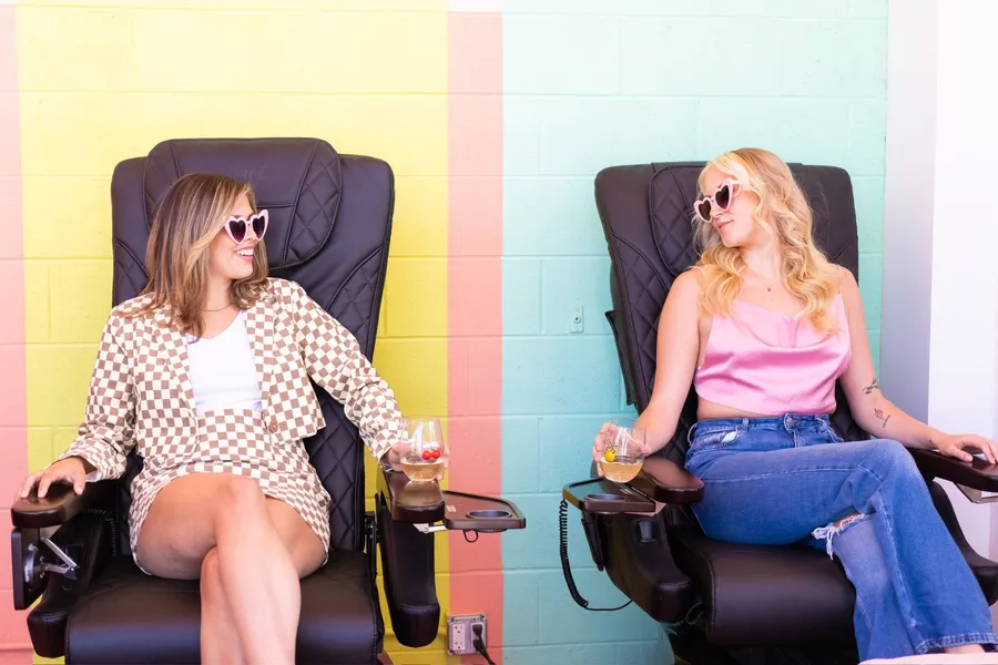 Two friends in heart sunglasses in pedicure chairs, in front of a pink, yellow and blue wall
